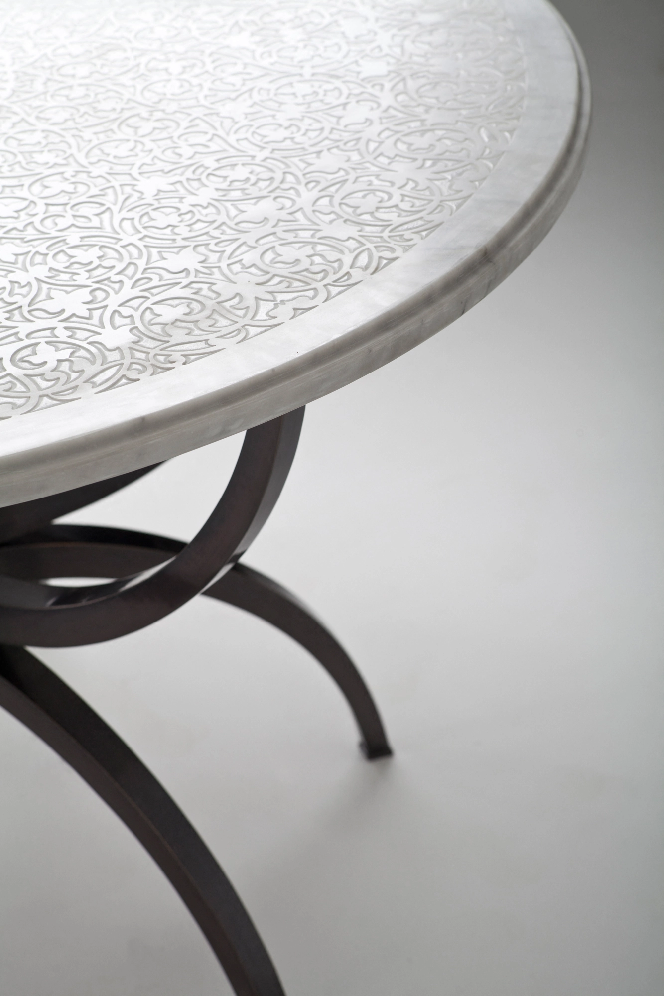 A round engraved pale-top table on curved dark iron legs, seen at an angle against a grey background.