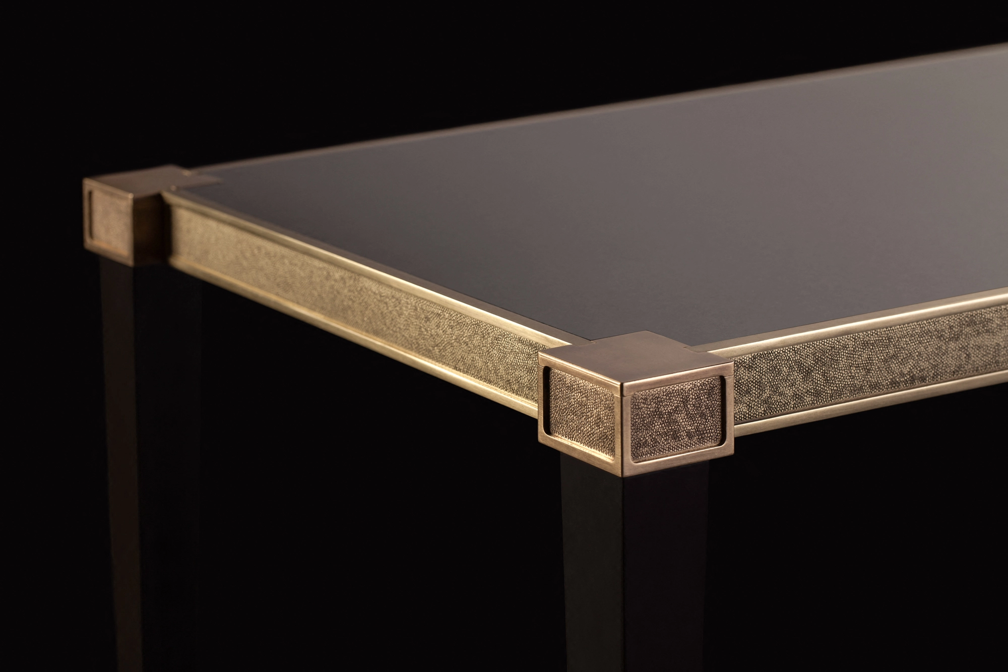 Corner detail of a console table with a dark top and a brass corner bracket, against a black background.