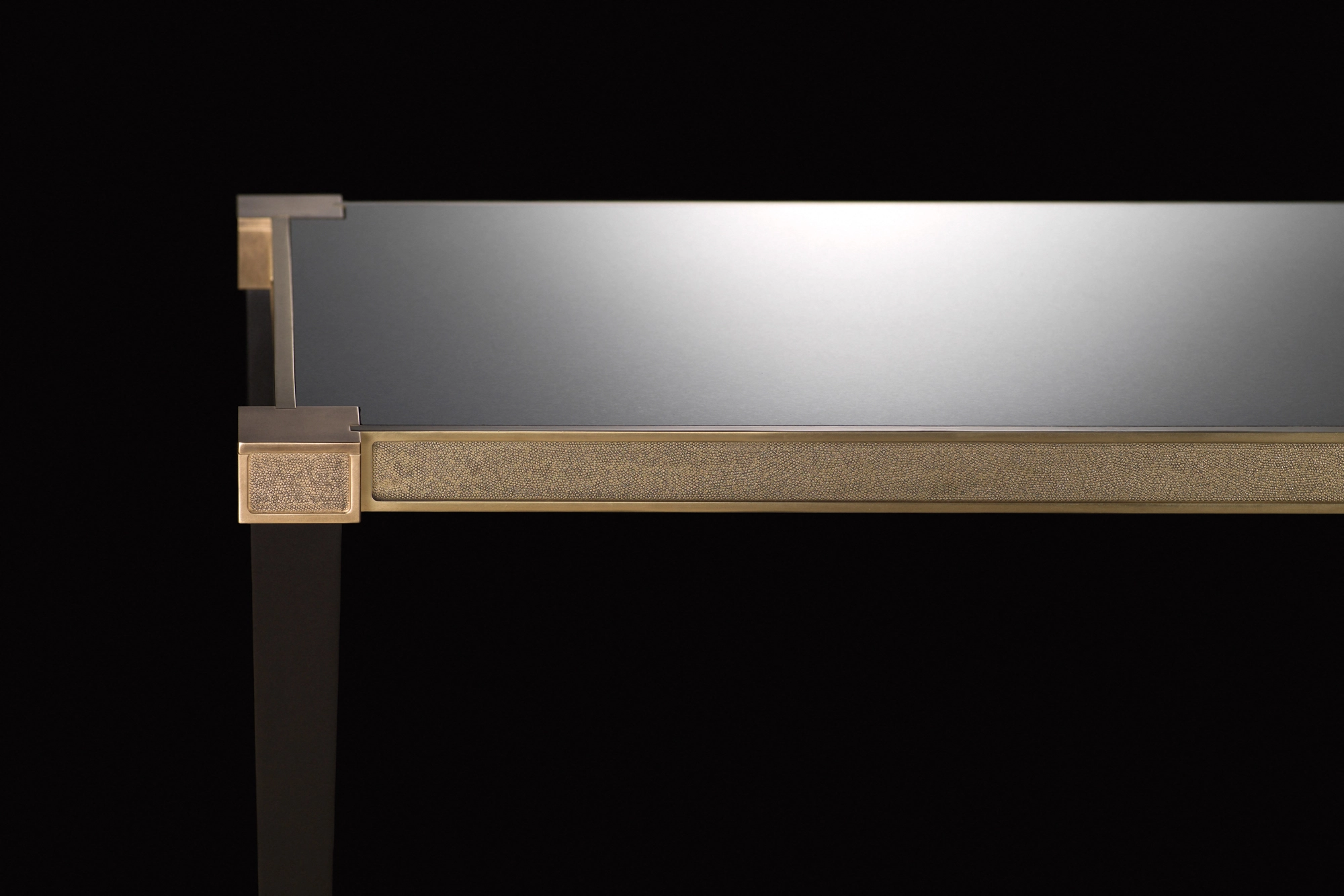 Corner detail of a console table with a mirrored top and brass edge, against a black background.