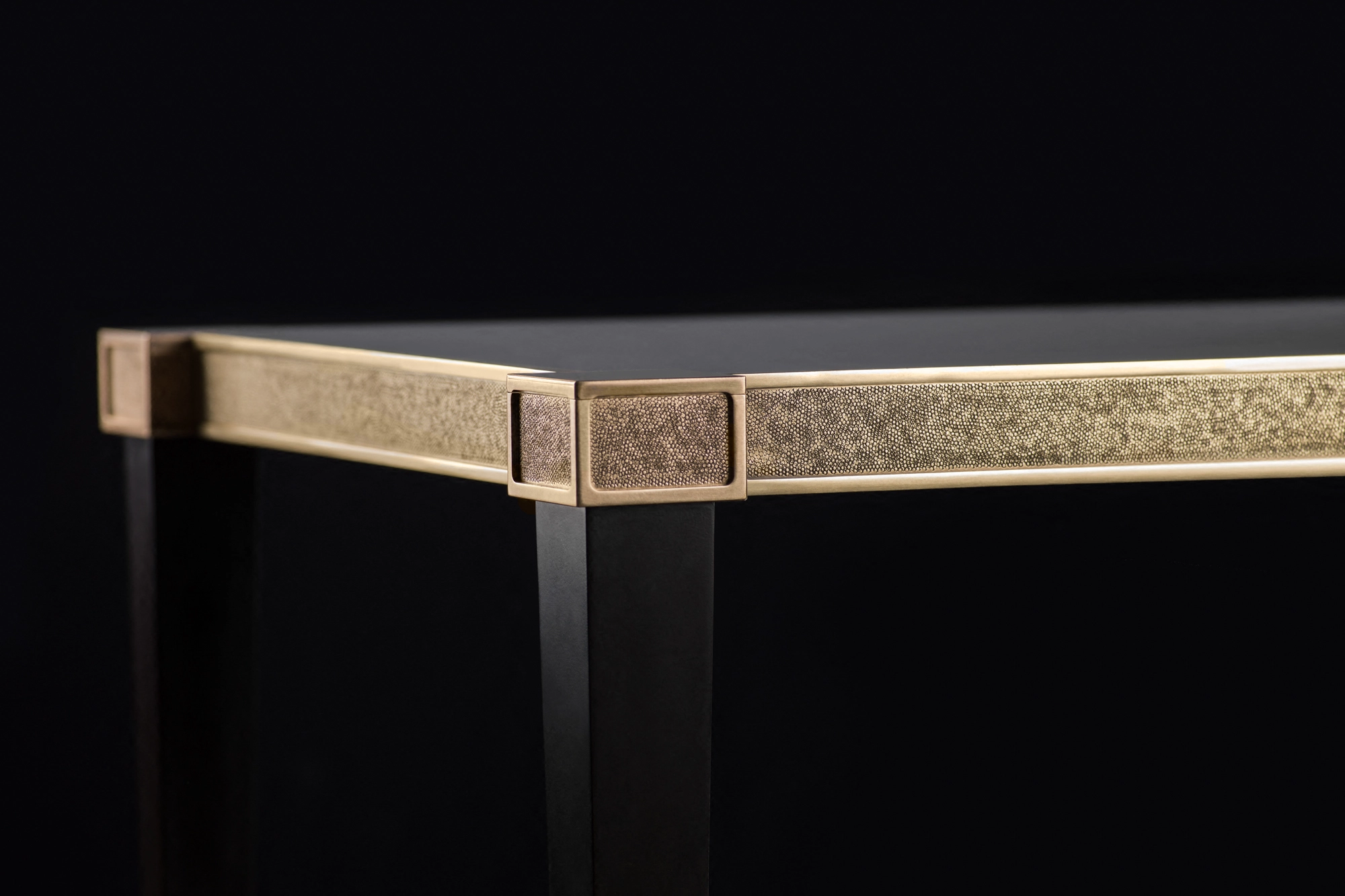 Corner of a console table with a dark top, brass trim and a textured side panel, on a black background.