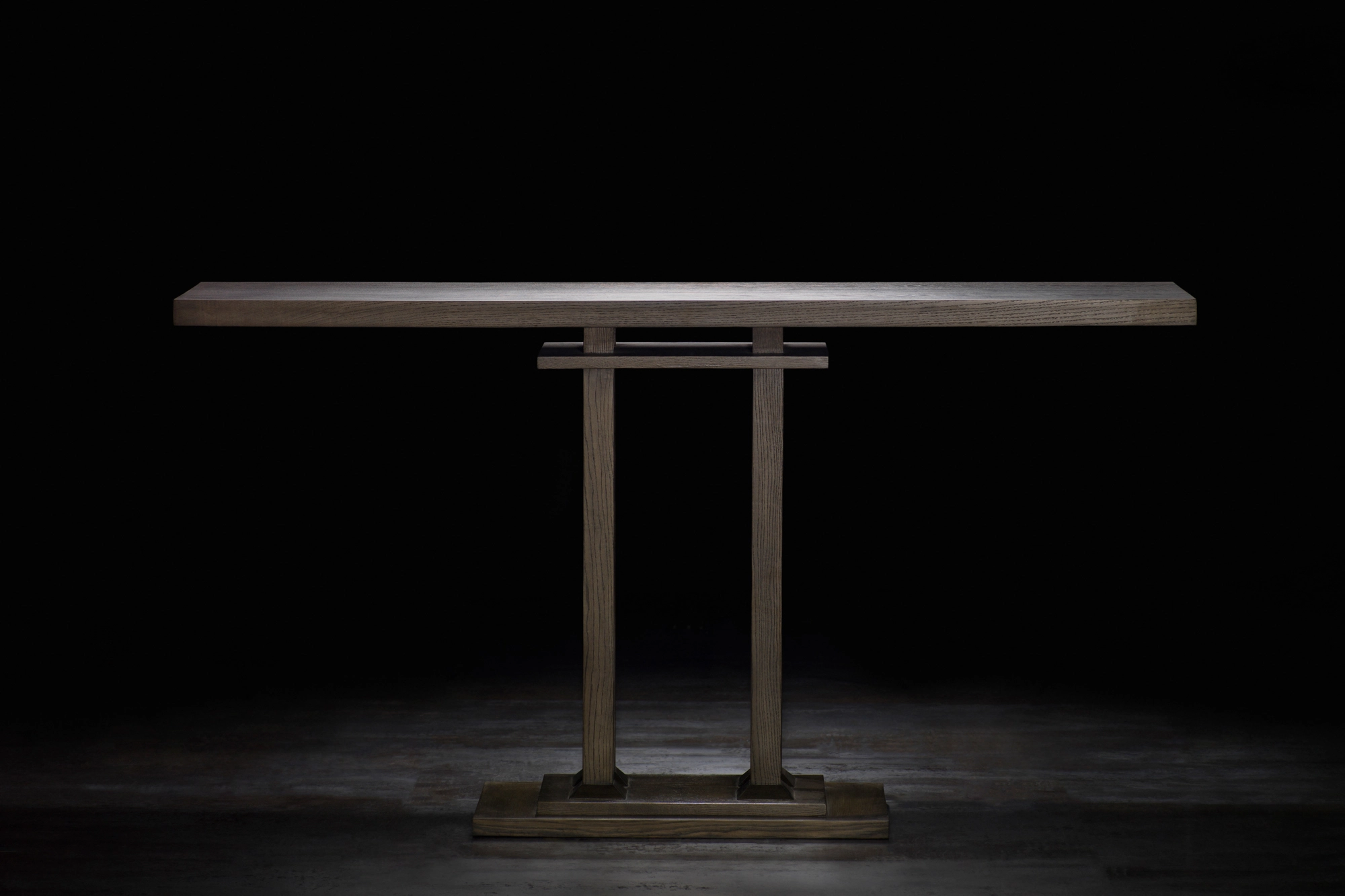 A wooden console table on a twin-column pedestal base, spotlit against a dark background.