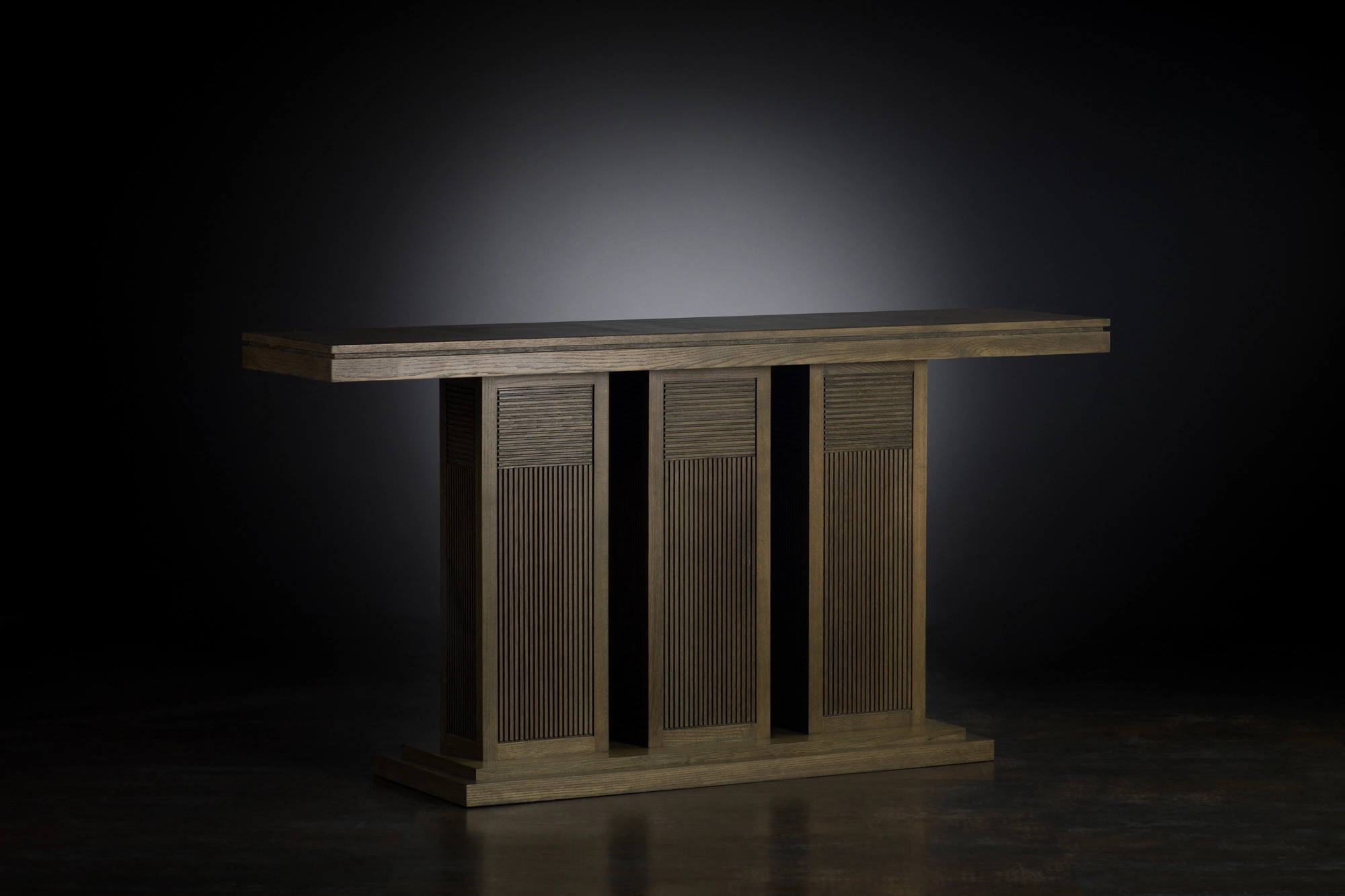 A wooden console table on a ribbed three-column base, spotlit against a dark background.