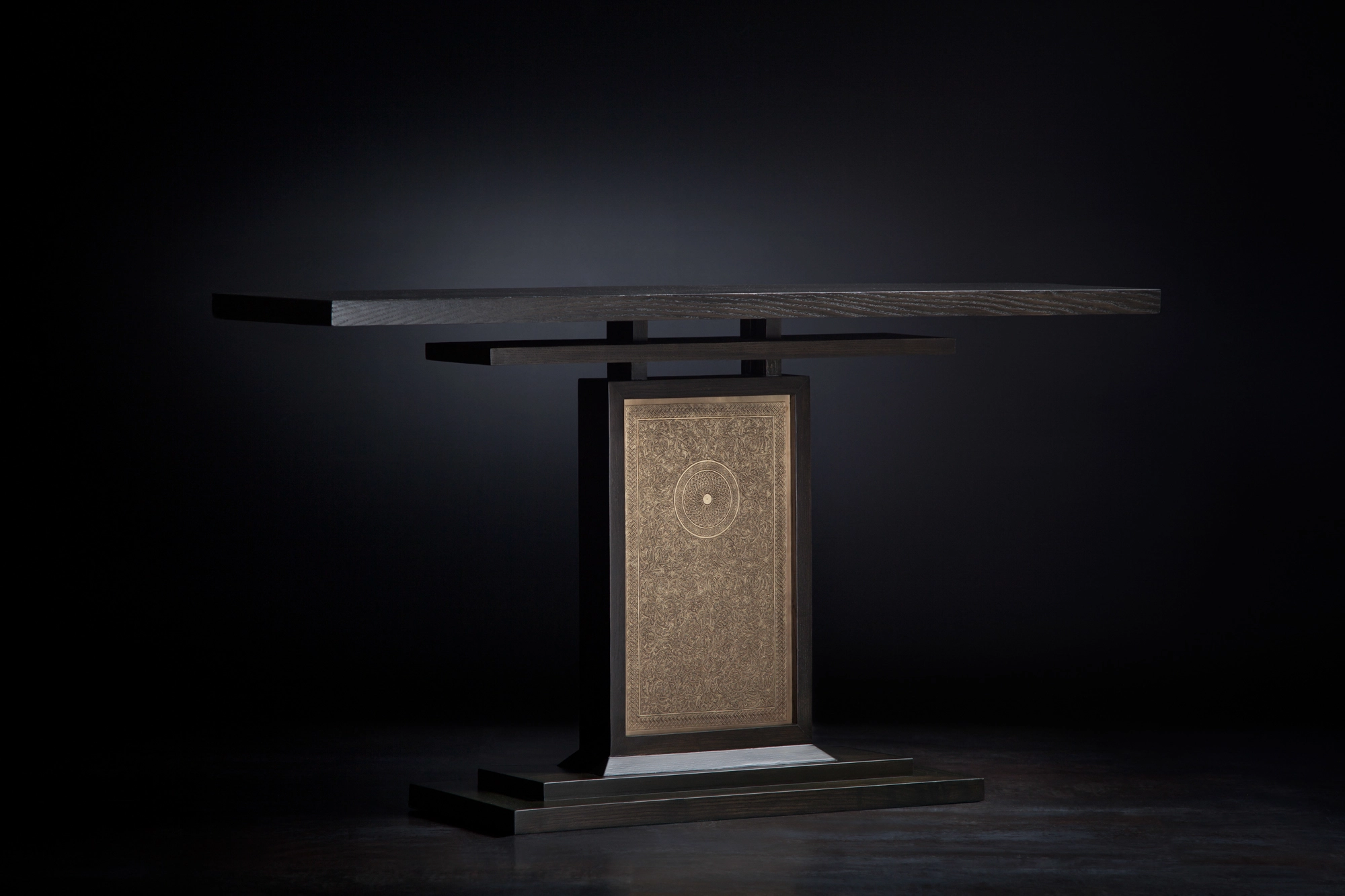 A console table with a central pedestal set with a pale engraved panel, lit against a dark background.