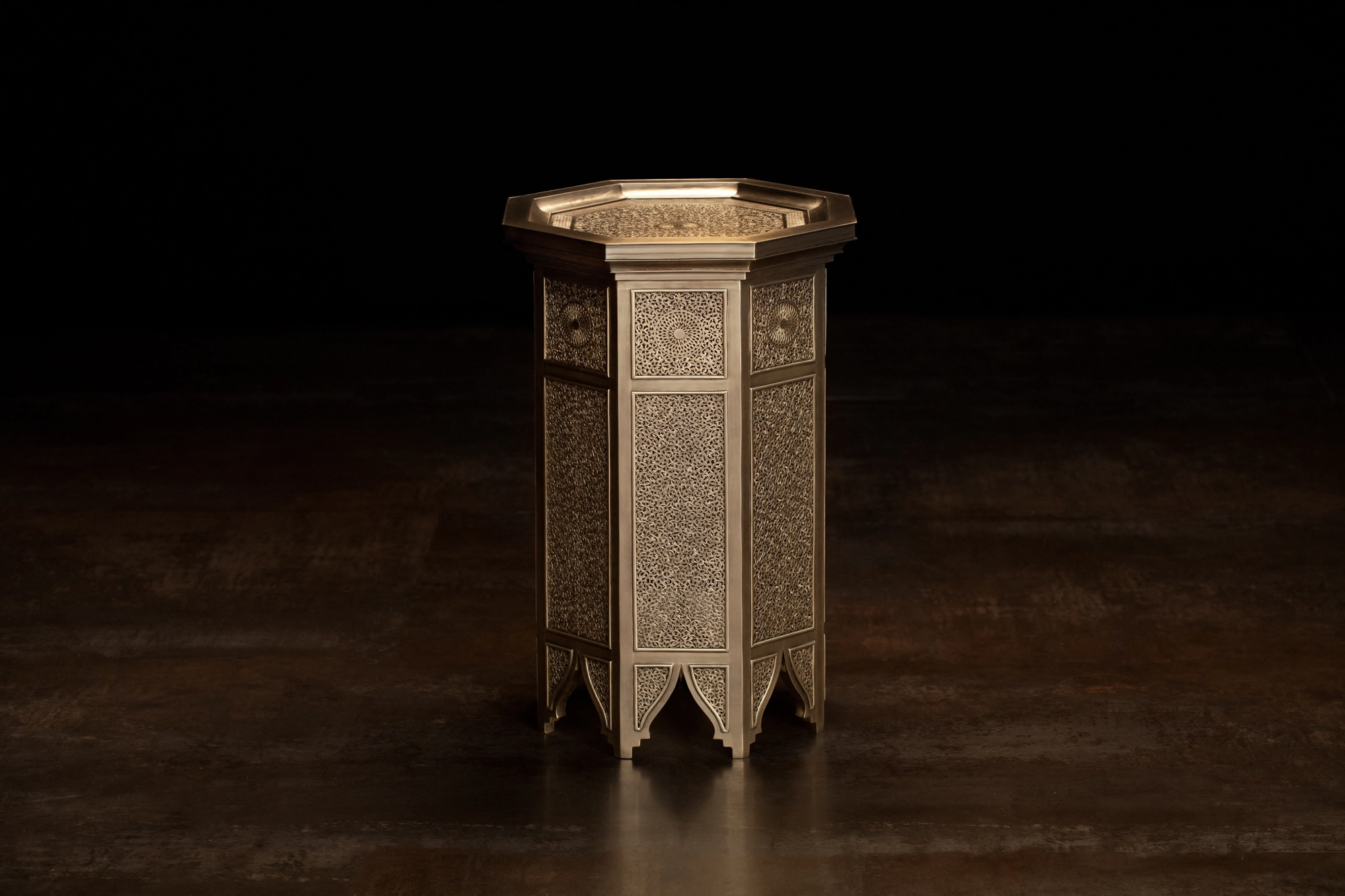 An octagonal engraved brass occasional table with arched feet, glowing warmly against a dark background.