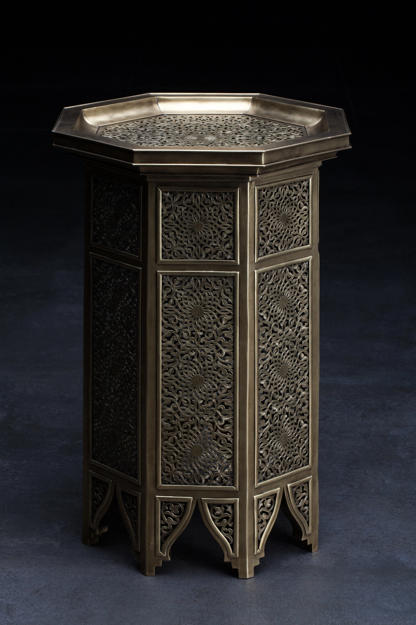An octagonal engraved brass occasional table with a tray top, seen at an angle against a dark background.