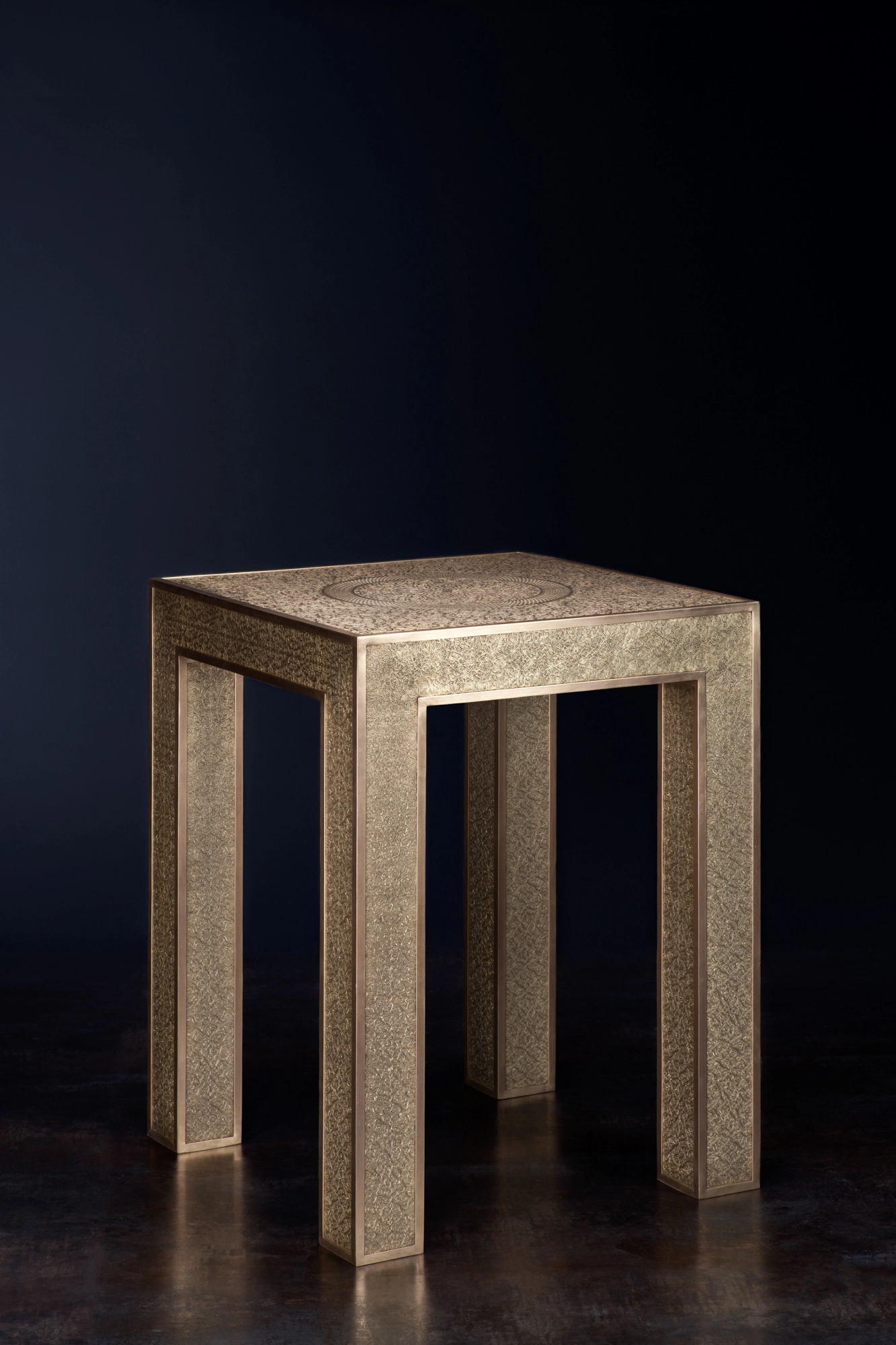 A small square engraved-metal occasional table on tapered legs, against a black background.