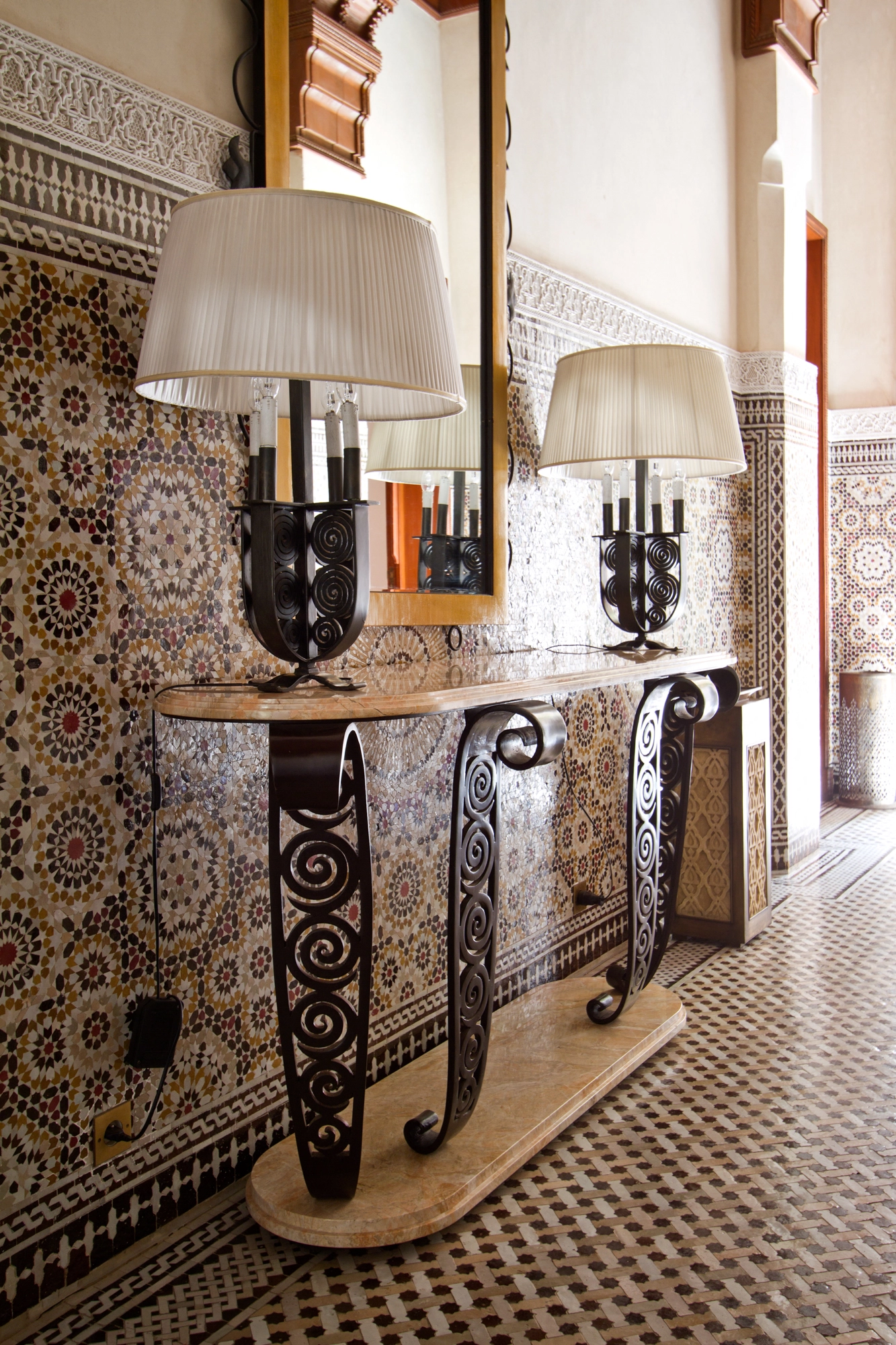 A console with ornate scrolled iron legs and a stone top holding table lamps, against a zellige-tiled wall.