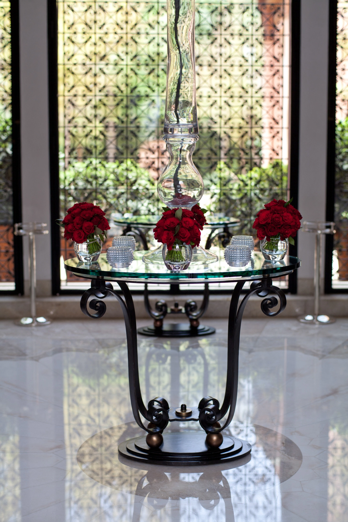 A round glass table on an ornate iron scroll base holding red roses and a candelabra, before a window.