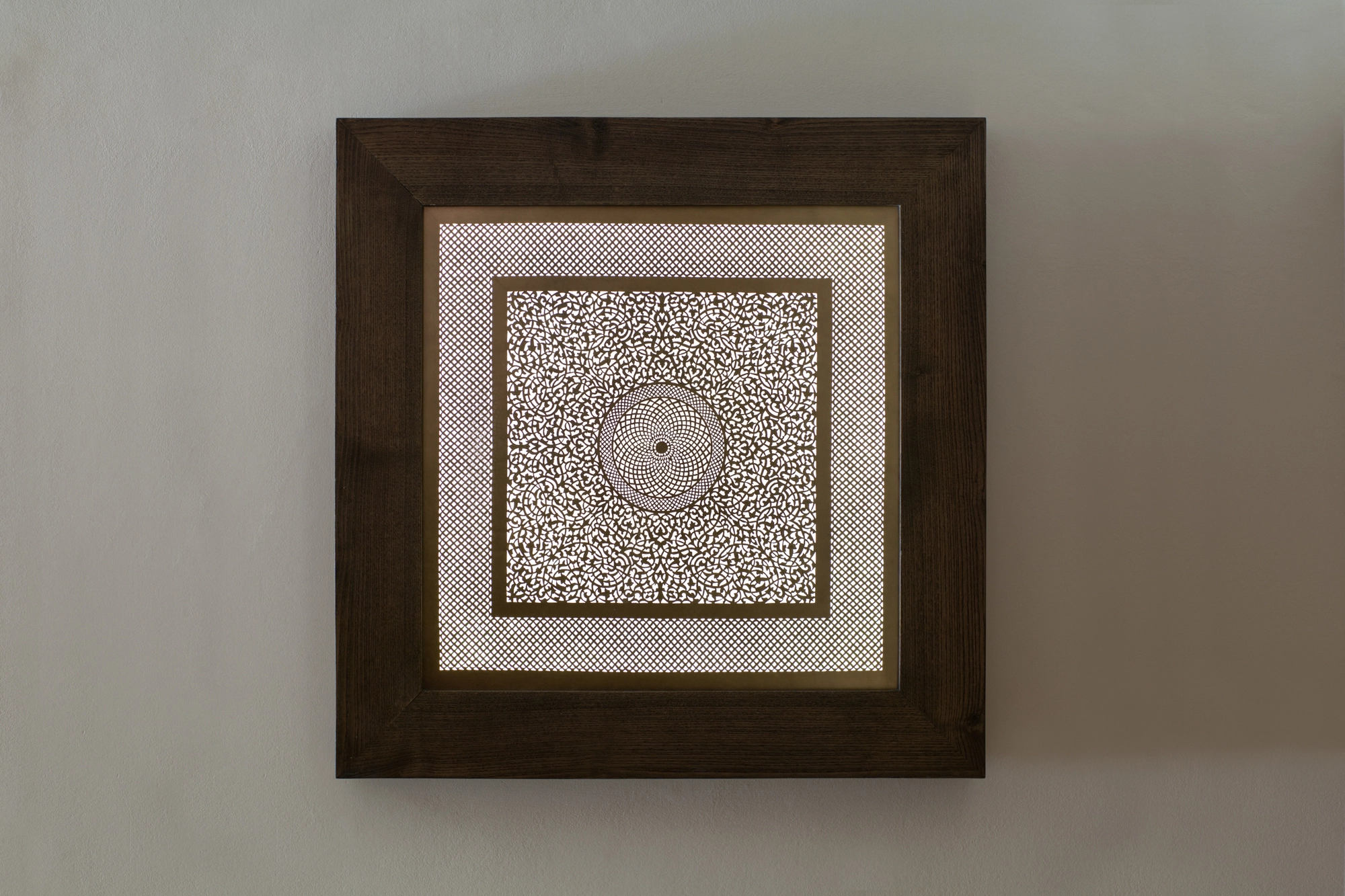 A small square framed panel with a pale pierced pattern and central medallion on a white wall.