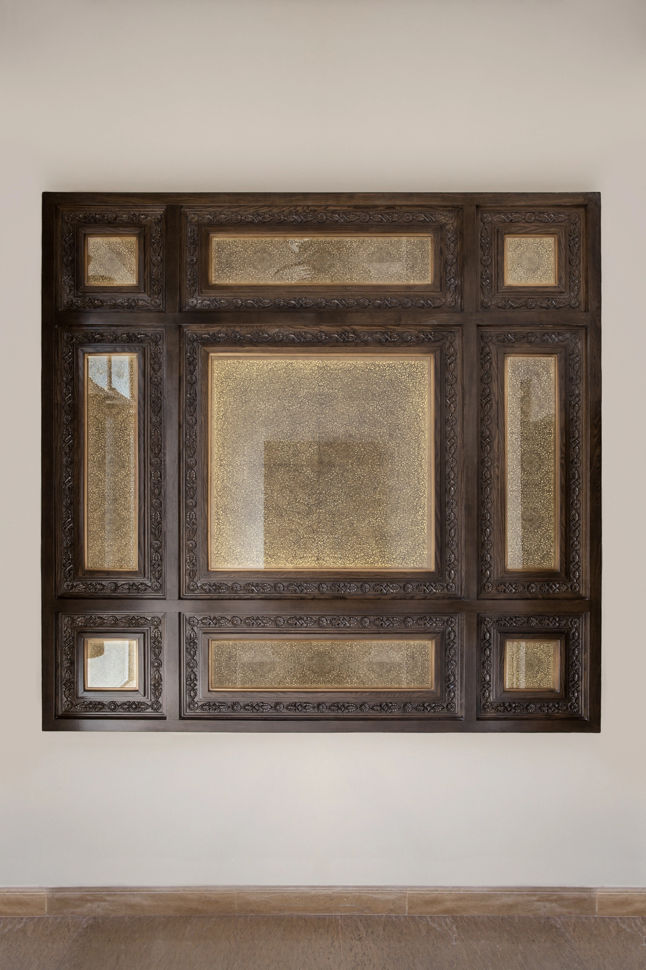 A large square wall panel of framed mirrored sections set in a carved dark wood surround on a white wall.