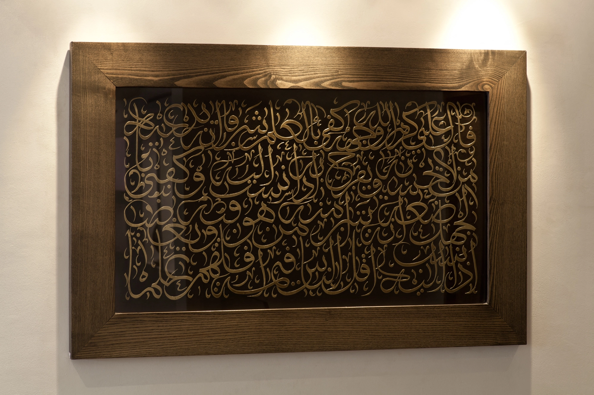 A framed panel of dark engraved Arabic calligraphy on a wall, lit from above.