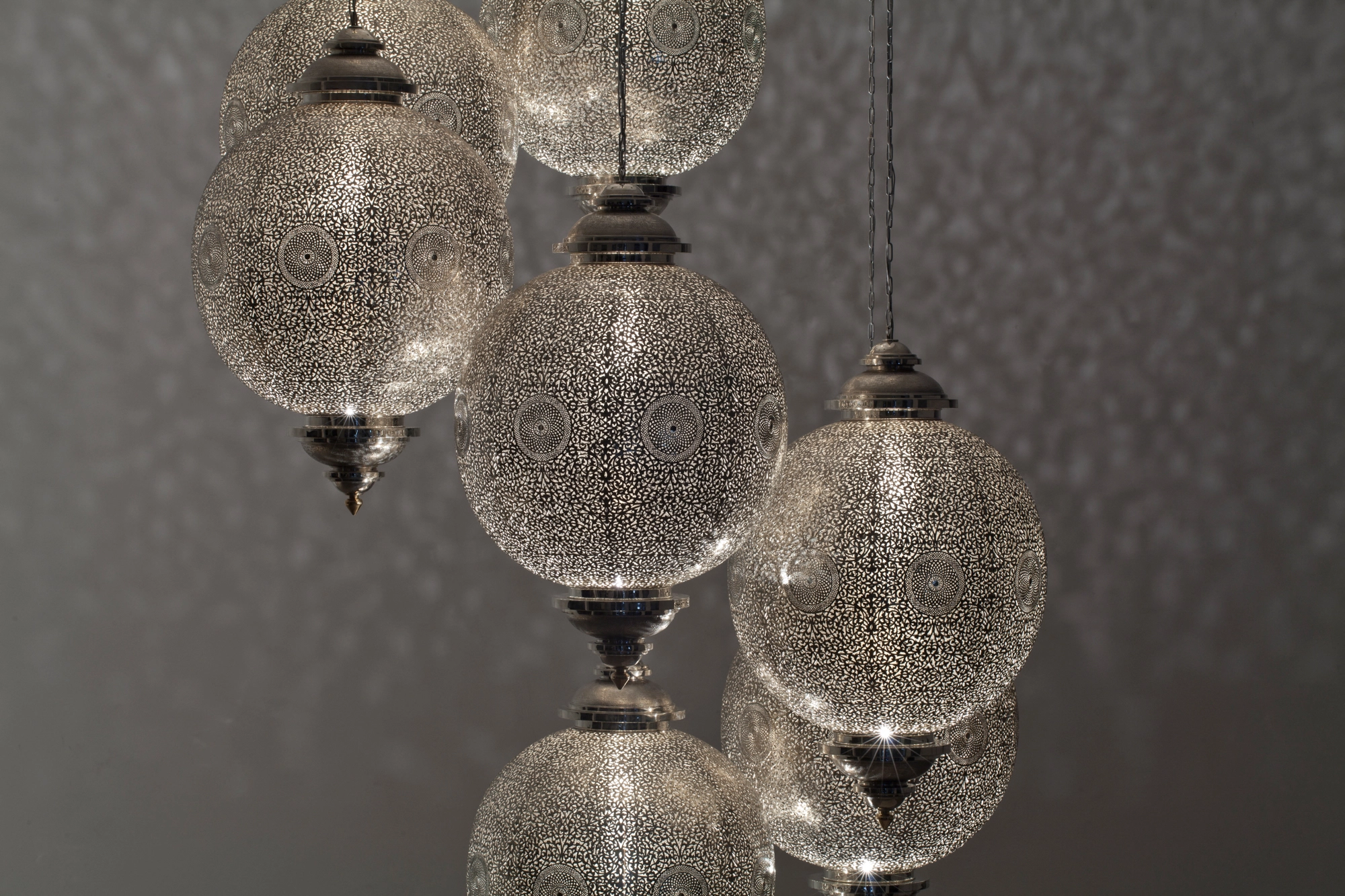 A cluster of pierced silver globe pendant lights suspended at staggered heights against a grey background.