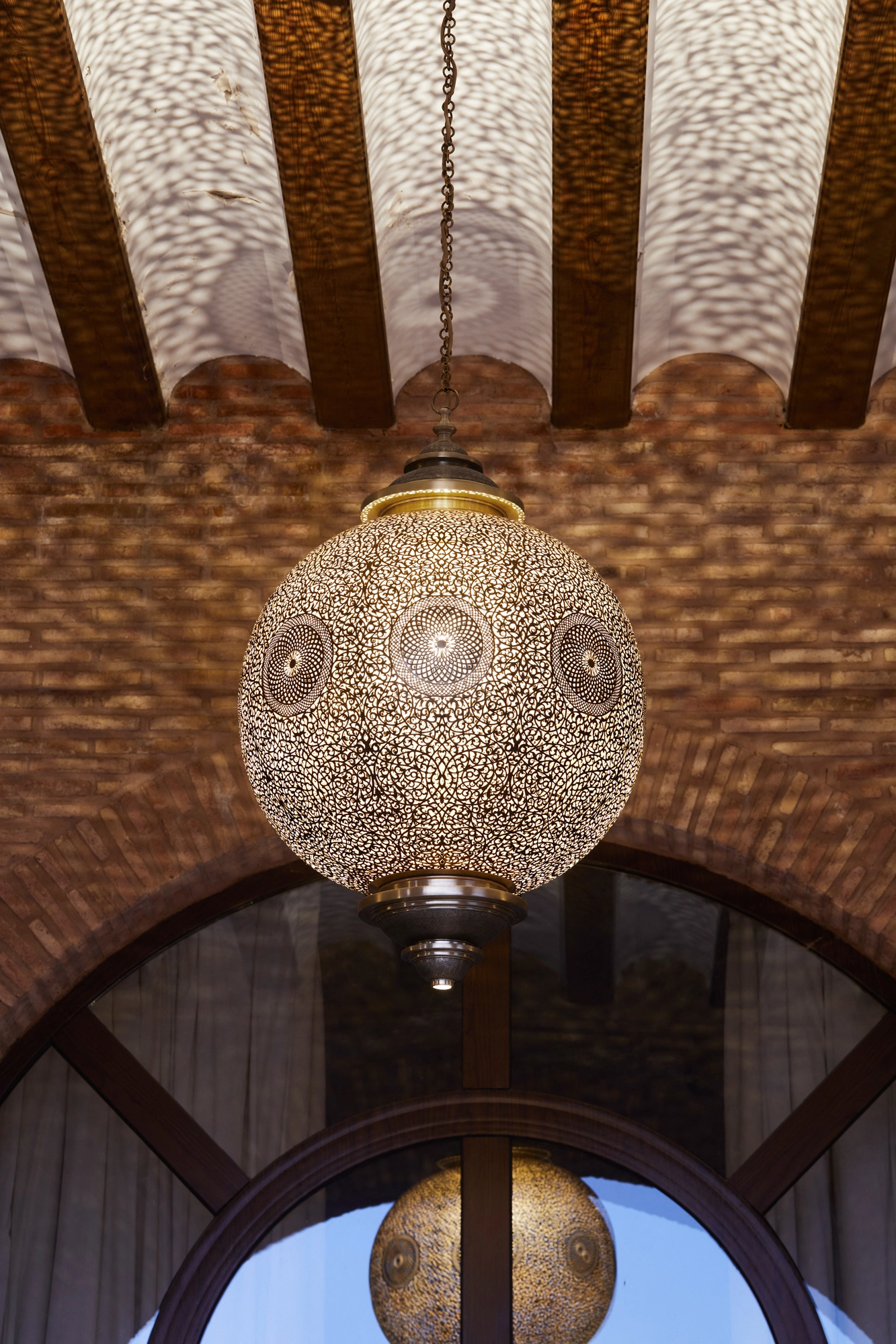 Pierced brass globe pendant lantern hanging beneath a vaulted ceiling of radiating wooden beams.