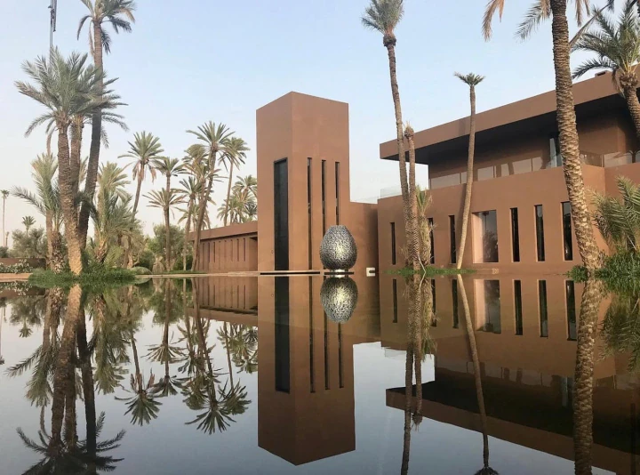 Modern villa mirrored in a pool with palms and a pierced metal sphere beside the water at dusk.
