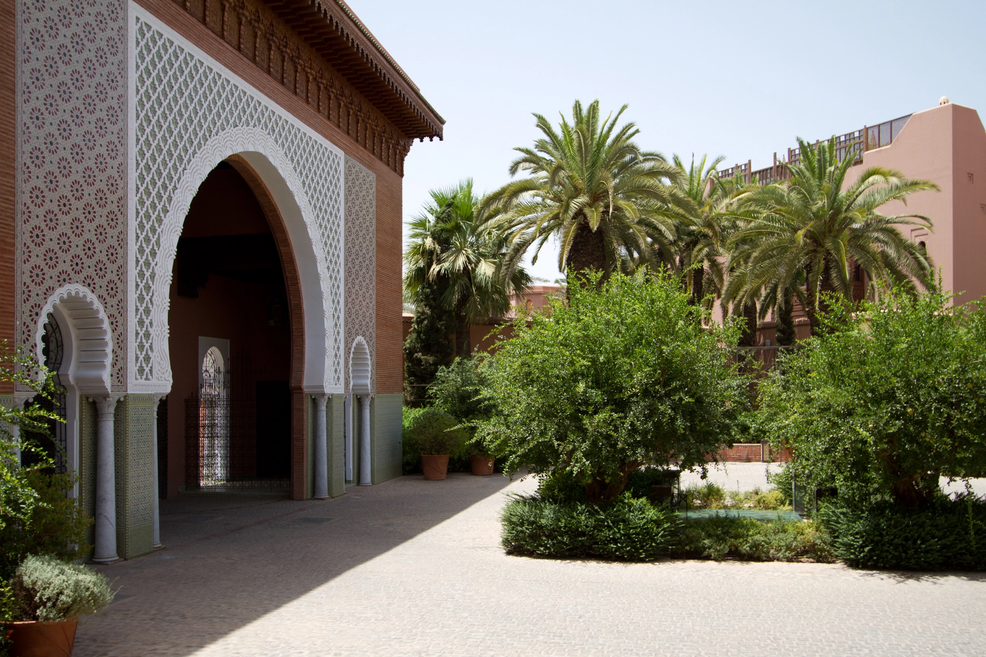 Sunlit Moroccan courtyard with a tall arched doorway framed by palms and clipped garden hedges.