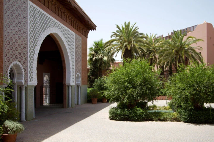 Sunlit Moroccan courtyard with a tall arched doorway framed by palms and clipped garden hedges.