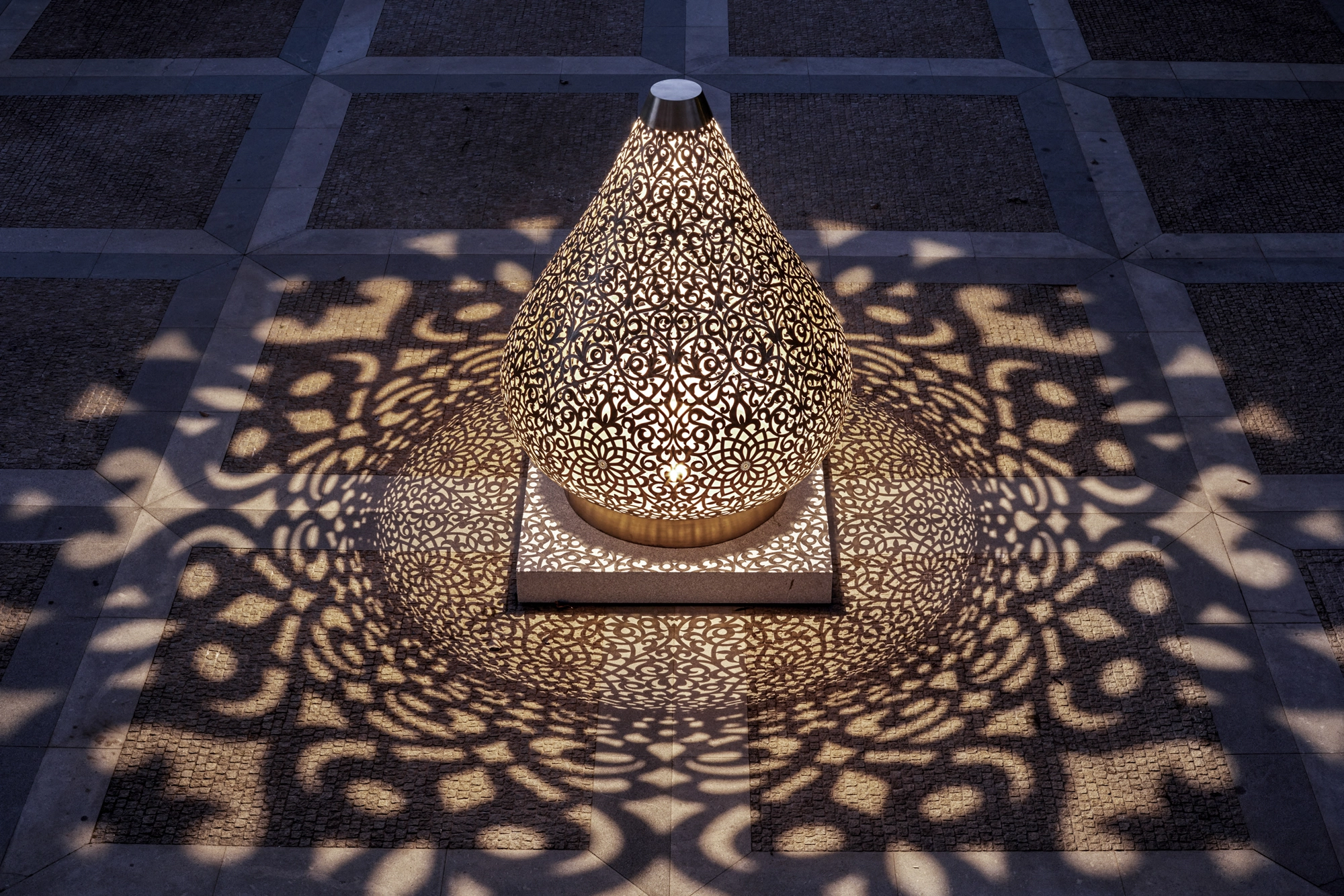 Teardrop-shaped pierced-metal Floral Lotus sculpture on a dark tiled floor, casting floral shadow patterns.